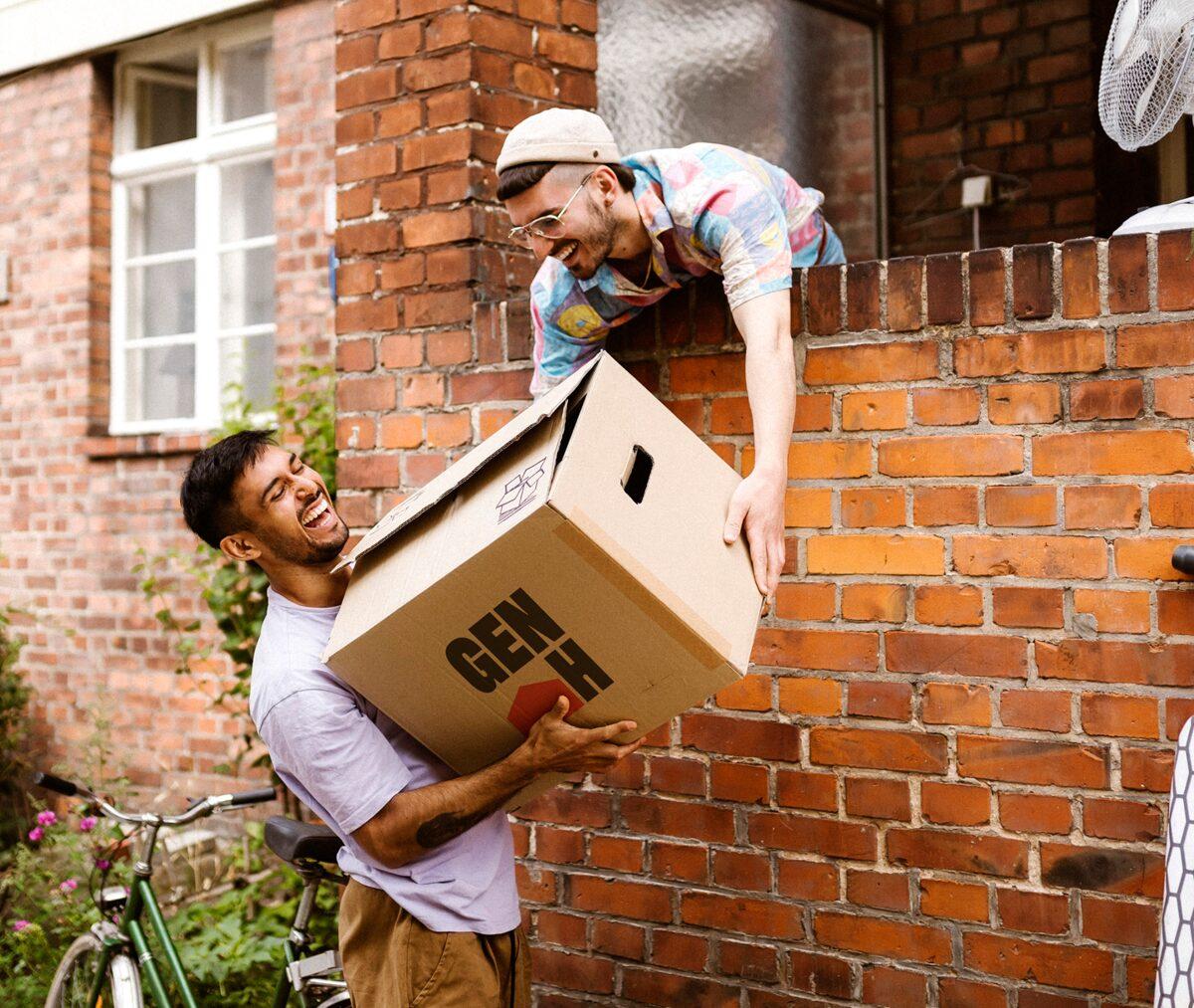 Two people passing a Gen H moving box over a garden wall